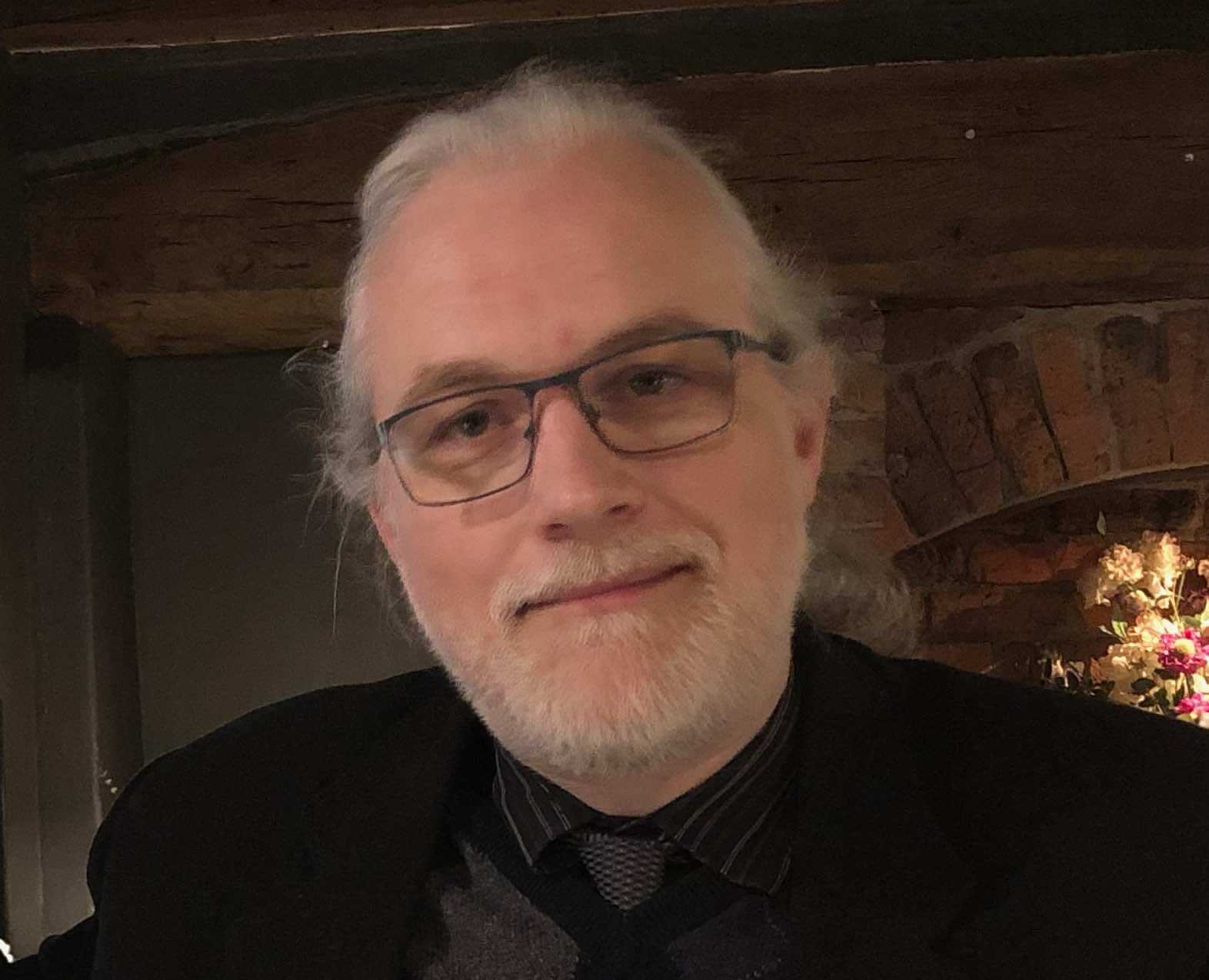 Martijn Dekker; a middle aged white man with glasses, grey hair and beard. He is dressed smartly in black suit and tie.